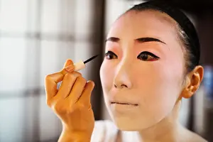 Mid Adult Women Prints: Geisha woman with traditional white face makeup painting on heavy eyeliner with a brush