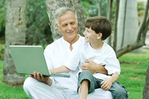 Affectionate Prints: Grandfather holding grandson on lap, boy looking at laptop computer, both smiling