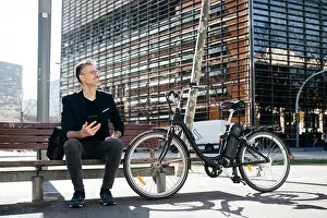 Environmental Issues Prints: Gray-haired businessman sitting on a bench next to electric bicycle in the city