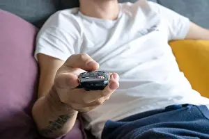 Lazy Prints: A thin guy in home clothes sits on a sofa and watches TV. Bored guy with tv remote control