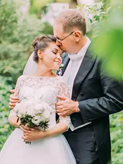 Newlywed Prints: Half-length outdoor portrait of the groom kissing the bride in teh forehead in the green garden