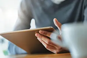 Young Person Prints: Hand of a Black man holding a tablet pc in close up viewed past a mug of coffee on a table