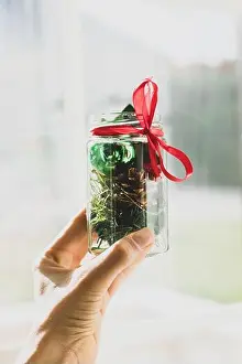 Presents Prints: Hand holding Christmas decorations in a jar with ribbon in front of a window