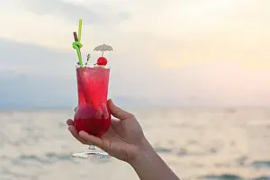 Cocktails Prints: Hand holding red cocktail drink in twilight sea & sky background