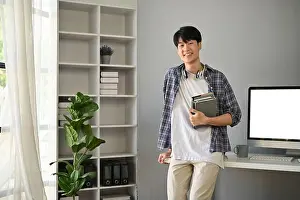 Developer Prints: A handsome and happy young Asian man in a flannel shirt stands in his modern office with books in his hand