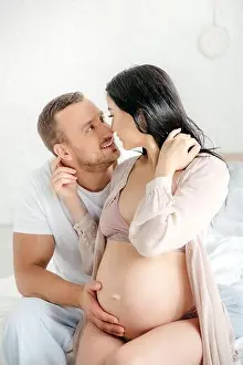 Closeness Prints: handsome smiling husband hugging and going to kissing his pregnant wife in bed