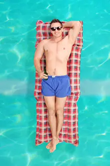 Mattress Prints: Handsome young man in blue swim trunks holding a bottle of beer and floating on water on a swim mattress