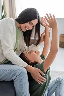Childcare Prints: happy brunette babysitter sitting on couch and looking at cheerful kid with outstretched hands