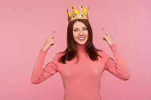 Toothy Smile Prints: Happy cheerful brunette woman in pink sweater pointing fingers on golden crown on her head