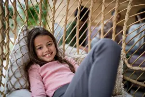 Look At Camera Prints: Happy little girl sitting in wicker rattan hang chair outdoors in patio in autumn