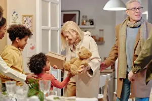 Brother Prints: Happy mature blond woman in beige coat passing teddybear to cute little grandson standing in front of her while meeting