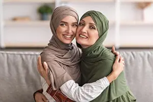 Middle Aged Prints: Happy Muslim Mother And Adult Daughter Hugging Wearing Hijab Indoor