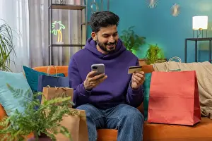 E Commerce Prints: Happy shopaholic consumer indian man sitting with shopping bags at modern home apartment making online order