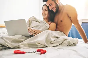 Happy young couple lying on a bed with computer - Beautiful married couple watching role games sex video on laptop laughing together