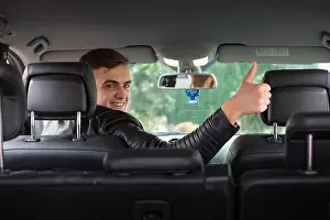 Hand Gesture Prints: Happy young man behind the steering wheel looking towards the back seat and giving a thumbs up