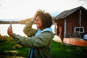Weekend Activities Prints: Happy young woman taking selfie with smart phone while standing against lake during sunset