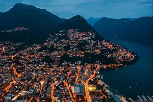 Aerial Cityscape Prints: High angle aerial drone night shot of city street lights by lake monte bre in Lugano