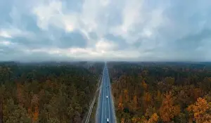 Highway road with passing cars in autumn forest with dark moody sky with clouds from aerial view