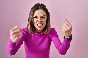 Hispanic woman standing over pink background angry and mad raising fists frustrated