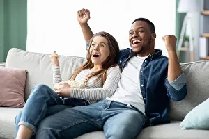 Multicultural Prints: Home Entertainment. Happy Interracial Couple Watching Tv And Cheering Sports At Home