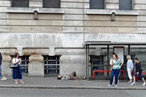 Asleep Prints: Homeless man sleeping on the ground next to a London bus shelter being ignored by waiting people outside Waterloo