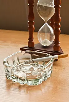 Nicotine Prints: Hourglass and smoking a cigarette in a glass ashtray on wooden top