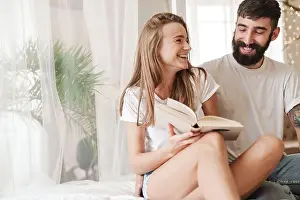 Optimistic Prints: Image of happy caucasian couple laughing and reading book while sitting on bed in bright room at home