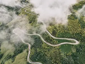 image taken with a drone of a long road with many curves on the slope of the green mountain between grassy vegetation