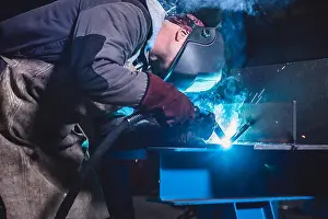 Place Of Work Prints: Industrial Welder With Torch and Protective Helmet in big hall welding metal profiles
