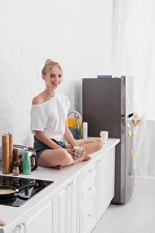 Housewife Prints: joyful woman sitting on kitchen table with crossed legs and looking at camera