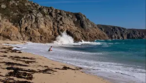 Beach Holiday Prints: One lady sitting on the sand with theSea swirling up the beach around her in waves that crash against the cliffs