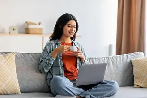 Full length of beautiful young Arab woman sitting cross legged on couch, working on laptop, drinking coffee at home