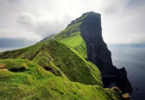 Kallur Lighthouse Prints: lighthouse sitting on top of a grassy hill close to a rocky black cliff looks over the tranquil sea on cloudy day