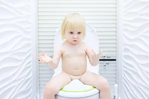 A little blonde girl with a mischievous look sits on the toilet in a brightly lit toilet room