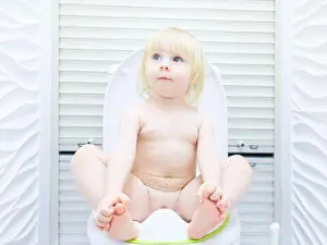 A little blonde girl with a mischievous look sits on the toilet in a brightly lit toilet room