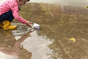 Rainy Prints: a little girl in a pink sweater and rubber boots launches a paper boat in a large puddle