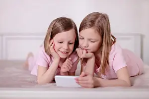 Pajamas Prints: Two little girls (sisters) in pink pajamas on the bed watching a smartphone and reading an e-book