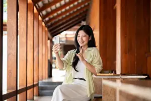 Asians Prints: Long hair asian woman talking a selfie on her phone while sitting at table in wooden house corridor