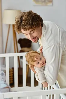 Curly Hair Prints: loving curly father lifting his infant son with blue eyes from baby crib in nursery