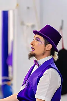 Magician wearing purple suit and top hat looking up with purple makeup on face and small goatee