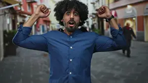 Wristband Prints: Man with curly hair raising arms on a cobbled city street with mouth open in surprise; surprise celebration