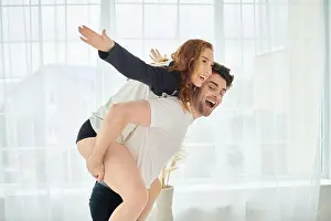A man effortlessly lifts a woman onto his back, showcasing strength and love in a beautiful bedroom setting