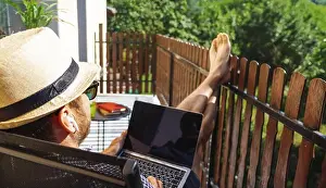 Well Being Prints: Man in hat and sunglasses listening to music with his headphones and using his laptop on the balcony