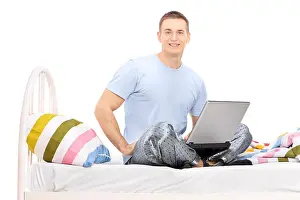 Pajamas Prints: Man in pajamas working on a laptop seated on a bed isolated on white background