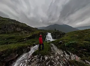 Red Jacket Prints: man in red stands under waterfall on black rock in mountains on cloudy rainy day