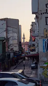 Minaret Prints: Man rides a scooter in the middle of a narrow run down street at sunset with a minaret visible behind