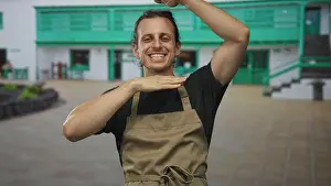 Lean Prints: Man smiling with hands framing face at building entrance wearing apron and showing forearms; welcoming joy