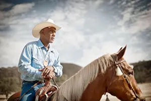 Checked Prints: Man wearing a cowboy hat sits on a horse resting his hands on the pommel and looks away with a watchful gaze
