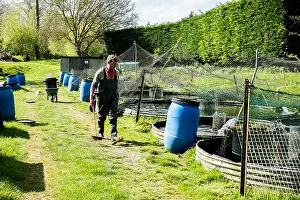 Mature Men Prints: Man wearing waders walking past water tanks at a fish farm raising trout
