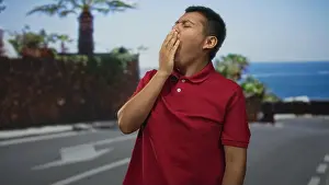 Fatigue Prints: Man yawning with hand covering mouth on seaside street wearing red polo shirt